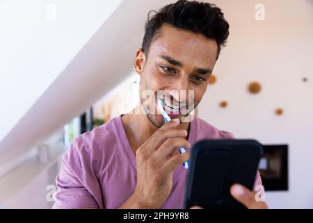 Heureux biracial homme se brossant les dents et en utilisant le smartphone dans la salle de bains Banque D'Images