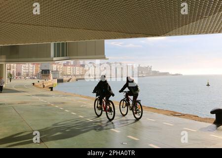 Deux cyclistes passant sous le Centre Botin à côté de la baie Santander Cantabria Espagne une matinée ensoleillée de printemps avec le Palais des fêtes au loin Banque D'Images