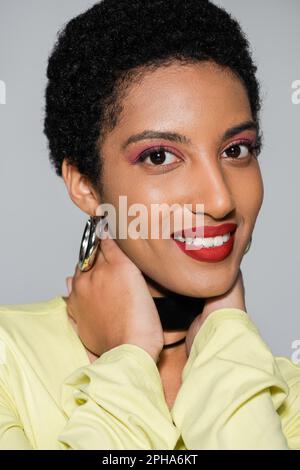 Femme afro-américaine insouciante avec maquillage touchant le cou isolé sur gris, image de stock Banque D'Images