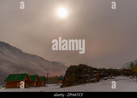 Chalets au bord de la rivière près de la forêt sur la neige sous la montagne avec brouillard et neige blanche, avec la lumière de la lune sur une nuit lumineuse à Altai in Banque D'Images