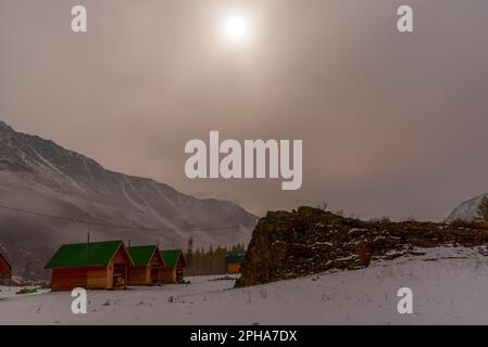 Chalets de vacances au bord de la rivière près de la forêt sur la neige sous la montagne avec brouillard et neige blanche, avec la lumière de la lune sur une nuit lumineuse dedans Banque D'Images