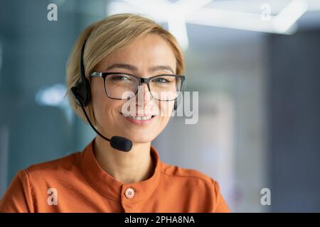 Portrait en gros plan d'une jeune belle femme blonde sur le lieu de travail, d'un employé du centre d'appels souriant et regardant la caméra, d'un opérateur utilisant un casque pour les appels vidéo et d'un service de consultation en ligne avec la clientèle. Banque D'Images