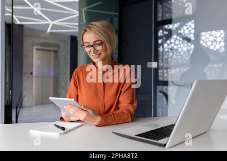 Jeune femme d'affaires prospère sur le lieu de travail est satisfaite des résultats de l'accomplissement au travail souriant et utilisant un ordinateur de tablette, blond programmeur teste de nouveaux logiciels dans l'application. Banque D'Images