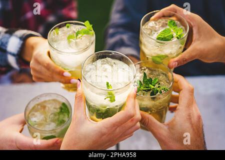 Gros plan sur les mains et les lunettes Mojito en cours de toasting dans un bar en plein air, avec le visage d'amis hors de vue, célébrant dans une atmosphère chaleureuse et animée. Banque D'Images