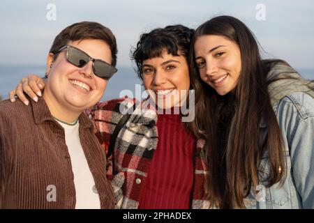 Trois amies féminines diverses embrassent et sourient au bord de la mer - une non binaire plus-taille avec des lunettes de soleil, cheveux courts, brunette avec cheveux ondulés, et un autre Banque D'Images