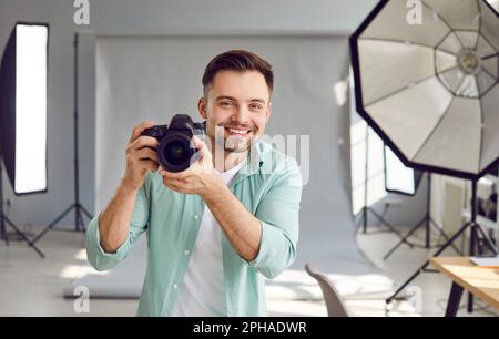 Un photographe souriant tenant un appareil photo debout dans un studio professionnel avec un équipement léger. Banque D'Images