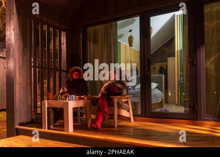 Une femme âgée dans un manteau et une fille dans un écossais sont assis dans la soirée sur la véranda près d'une maison de vacances en bois avec des portes vitrées. Banque D'Images