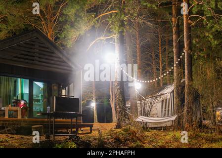 Un brazier avec feu et charbon de bois pour un barbecue se tient la nuit près d'un chalet de vacances avec une femme sur une chaise avec lanternes et guirlandes en Alt Banque D'Images