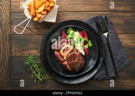 Steaks Osso Buco. Bœuf grillé Osso Buco ou steak de viande de portreuse avec garniture de salade et de frites sur un panneau de marbre noir sur du vieux bois Banque D'Images