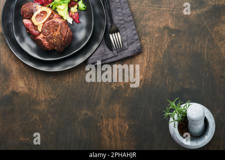 Steaks Osso Buco. Bœuf grillé Osso Buco ou steak de viande de portreuse avec garniture de salade et de frites sur un panneau de marbre noir sur du vieux bois Banque D'Images