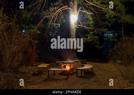 Un foyer, un foyer en métal entouré de bancs en bois, se dresse la nuit sous un arbre avec une lanterne et un feu sur la rive de la rivière. Banque D'Images