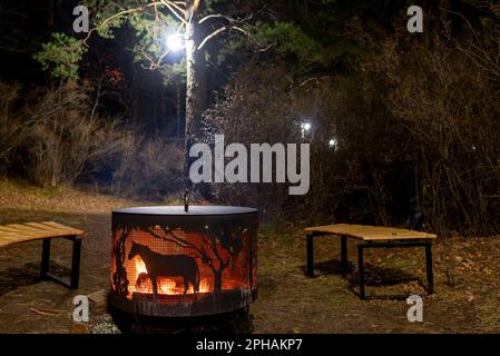 Un foyer, un foyer en métal, avec une silhouette de cheval, entouré de bancs en bois, se dresse la nuit sous un arbre avec une lanterne et un feu sur le Th Banque D'Images