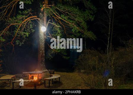 Un foyer, un foyer en métal entouré de bancs en bois se dresse la nuit sous un arbre avec une lanterne et un feu sur les rives de la rivière Katun. Banque D'Images