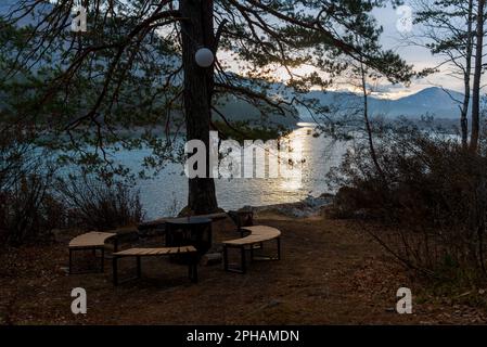 Un foyer, un foyer en métal entouré de bancs en bois se dresse à l'aube sous un arbre avec une lanterne sur la rive de la rivière Altai. Place pour le repos. Banque D'Images