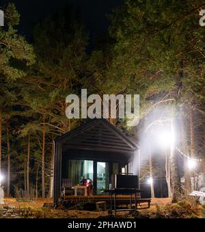 Un brazier avec feu et fumée pour un barbecue se tient la nuit près d'un chalet avec une fille avec un téléphone sur une chaise pour se détendre avec des portes en verre dans Un Banque D'Images