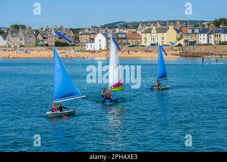 Les jeunes qui profitent du soleil d'été naviguent à bord d'un canot pneumatique dans le port d'Elie, avec la plage et la ville d'Elie en arrière-plan - Elie, Neuk est de Fife, Écosse, Royaume-Uni Banque D'Images