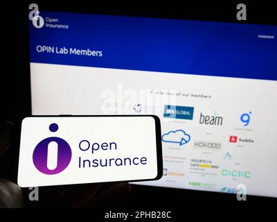 Personne tenant un téléphone portable avec logo de l'initiative de l'industrie Open Insurance à l'écran en face de la page Web. Mise au point sur l'affichage du téléphone. Banque D'Images