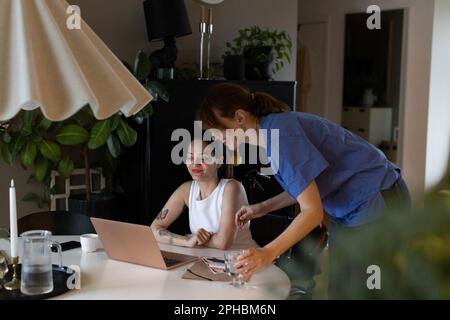 Une femme de soins parle à une femme avec un ordinateur portable de paraplégie à la maison Banque D'Images