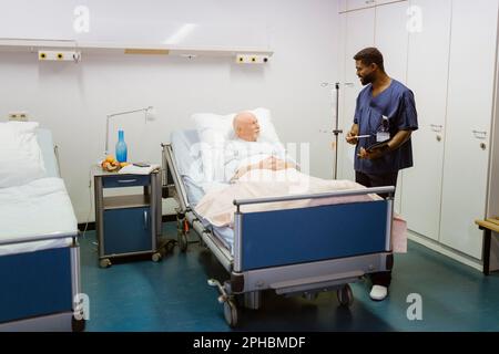 Infirmière de sexe masculin parlant à un patient âgé allongé sur le lit à l'hôpital Banque D'Images