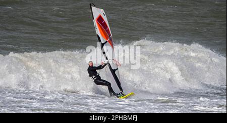 Planche à voile sportive en combinaison noire pratiquant la planche à voile classique le long de la côte de la mer du Nord par temps venteux pendant la tempête d'hiver Banque D'Images