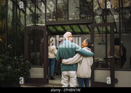 Vue arrière des amis hommes et femmes âgés debout les uns autour des autres lors du dîner Banque D'Images
