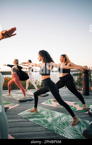 Des amies pratiquant le Warrior 2 posent sur le patio au coucher du soleil Banque D'Images