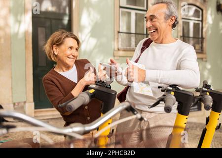 Les conjoints seniors louent des scooters électriques en utilisant le téléphone gesturant comme à l'extérieur Banque D'Images