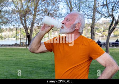 Un homme âgé en bonne santé prend son souffle après avoir fait de l'exercice dans le parc public de la ville au bord de la rivière. Eau potable d'une bouteille en plastique. Banque D'Images