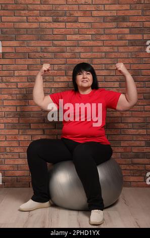 Bonne femme adulte en surpoids assise sur le ballon de fitness près du mur de briques à l'intérieur Banque D'Images