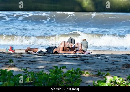 Jeune couple regardant leur téléphone cellulaire en étant allongé sur la plage à Palm Cove, Cairns Northern Beaches, Far North Queensland, FNQ, QLD, Australie Banque D'Images