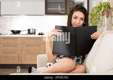 Jeune femme sur son salon canapé charmant sourire, peeking de derrière un catalogue noir ou un bloc-notes. L'arrière-plan flou présente un espace moderne Banque D'Images