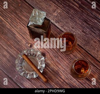 Cigare cubain dans un cendrier en cristal, une bouteille et deux verres de whisky sur une table en bois. Vue de dessus, plan d'agencement. Banque D'Images