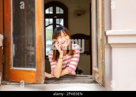 une étudiante portant un élégant manteau gris discutant au téléphone tout en lisant un livre, assise sur un banc de bois sur le campus universitaire. Banque D'Images