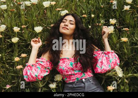 De dessus de jeune femme souriante avec de longs cheveux foncés et les mains levées couché sur des plantes vertes avec fleurs blanches en pleine lumière du soleil Banque D'Images
