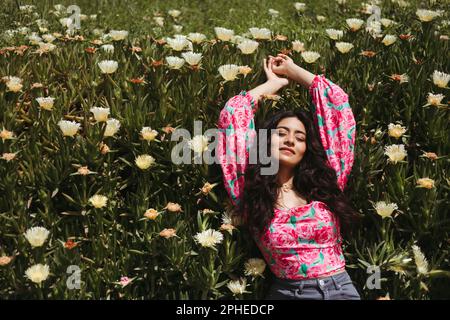 De dessus de jeune femme souriante avec de longs cheveux foncés et les mains levées couché sur des plantes vertes avec fleurs blanches en pleine lumière du soleil Banque D'Images