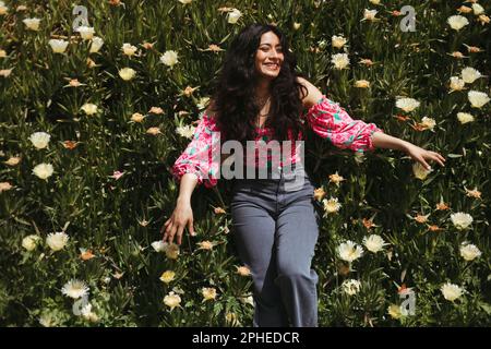 De dessus de jeune femme souriante avec de longs cheveux foncés regardant loin tout en étant allongé sur des plantes vertes avec fleurs blanches en pleine lumière du soleil Banque D'Images
