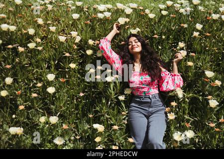 De dessus de jeune femme souriante avec de longs cheveux foncés et les mains levées couché sur des plantes vertes avec fleurs blanches en pleine lumière du soleil Banque D'Images