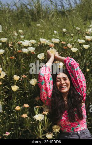 De dessus de jeune femme souriante avec de longs cheveux foncés et les mains levées couché sur des plantes vertes avec fleurs blanches en pleine lumière du soleil Banque D'Images