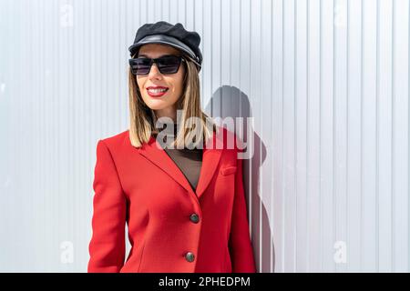 Jeune femme avec rouge à lèvres en manteau rouge et chapeau souriant et regardant l'appareil photo tout en se tenant contre le mur gris Banque D'Images