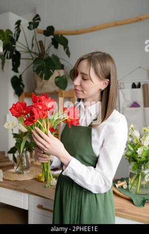 Jeune fleuriste femme souriante debout à table et tenant un bouquet de fleurs rouges fraîches tout en travaillant dans un studio de fleuriste Banque D'Images