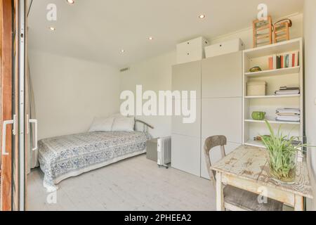 Intérieur de chambre contemporaine avec lit confortable placé près des étagères et table en bois avec plante en pot dans l'appartement entouré de murs blancs Banque D'Images