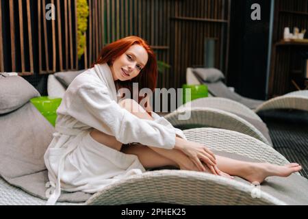 Portrait d'une jeune femme mignonne portant un peignoir blanc se relaxant sur une chaise longue au bord de la piscine après un soin de beauté au salon de spa, en regardant un appareil photo. Banque D'Images