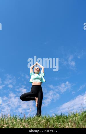 Femme faisant l'exercice de yoga Vrikshasana pose d'arbre dans la nature contre le ciel bleu. Banque D'Images