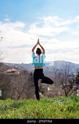 Vue arrière femme faisant l'exercice de yoga Vrikshasana pose d'arbre dans la nature contre la montagne. Banque D'Images