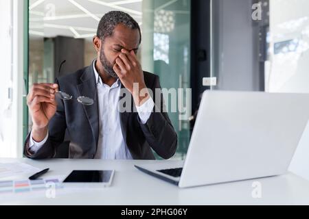 Un homme surtravaillé sur le lieu de travail à l'intérieur du bureau, un homme d'affaires afro-américain travaillant assis avec un ordinateur portable au travail, un patron qui délasse des lunettes et massante les yeux. Banque D'Images