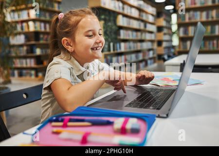 Étudiante gaie faisant ses devoirs à l'aide d'un ordinateur portable dans un club après l'école primaire. Retour à l'école. Enfant utilisant la technologie sur l'ordinateur élémentaire Banque D'Images