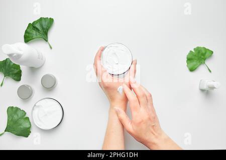 Femme appliquant de la crème hydratante cosmétique pour les mains. Produits cosmétiques, feuilles vertes sur table blanche. Spa, manucure, concept de soins de la peau. Pose à plat, en hauteur Banque D'Images