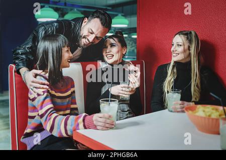 Trois amies de sexe féminin aiment prendre un cocktail dans un bar à thème de 90s, un ami souriant s'approche et encadre deux d'entre elles, tables de billard en arrière-plan. Banque D'Images