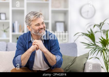 Portrait d'un bel homme effrayant aux cheveux gris dans une chemise bleue assise à la maison sur le canapé et regardant avec sourire sur le côté, croisant ses bras devant lui. Banque D'Images