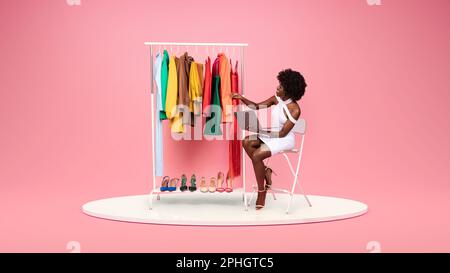 Souriante jeune femme afro-américaine designer de mode en robe blanche avec ordinateur portable regarde les vêtements Banque D'Images